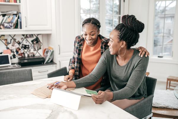 A black mother and her teenage daughter sit at a kitchen counter and smile while they look at an iPad